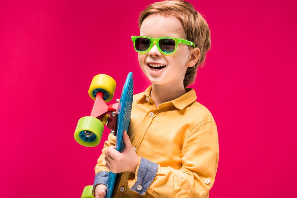 стильный улыбающийся мальчик в солнцезащитных очках позирует со скейтбордом изолирован на красный
 - Фото, изображение