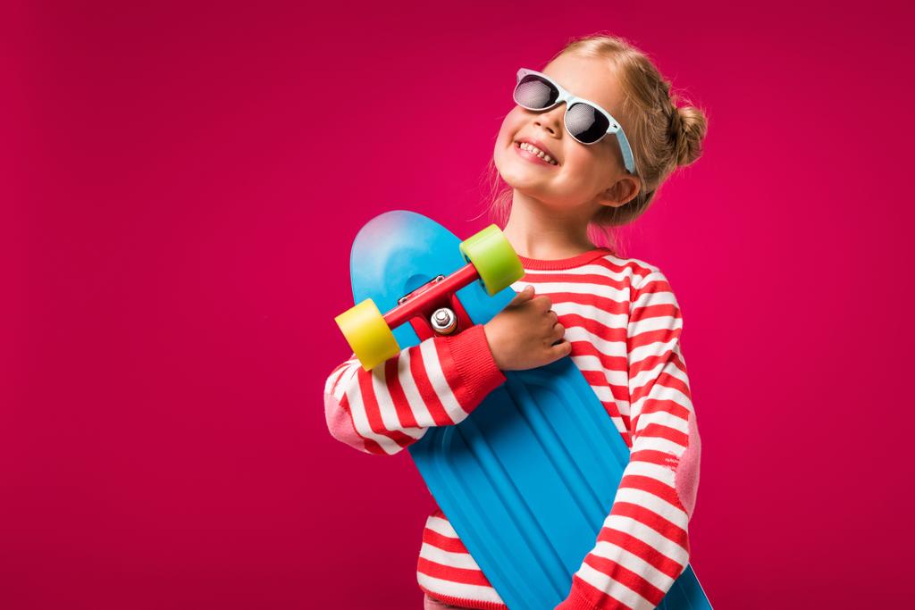 ευτυχισμένο παιδί κομψό σε γυαλιά ηλίου που θέτουν με skateboard που απομονώνονται σε κόκκινο - Φωτογραφία, εικόνα