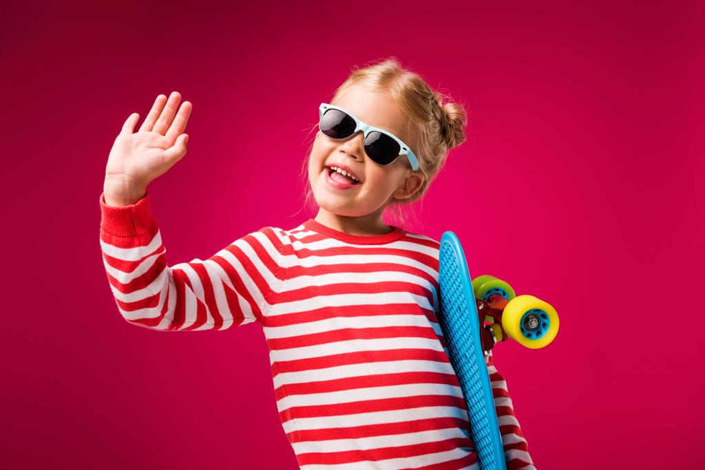 excited stylish kid in sunglasses holding penny board and waving isolated on red - Photo, Image