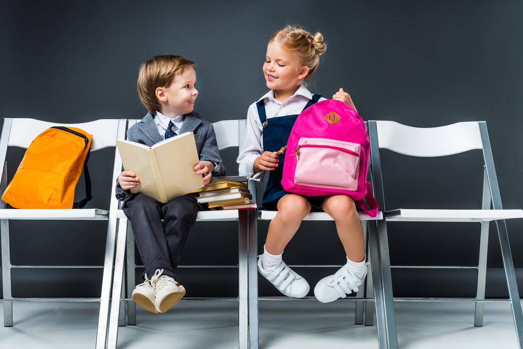 Улыбающиеся одноклассники сидят на стульях с книгами и рюкзаками, пока школьник читает книгу
 - Фото, изображение
