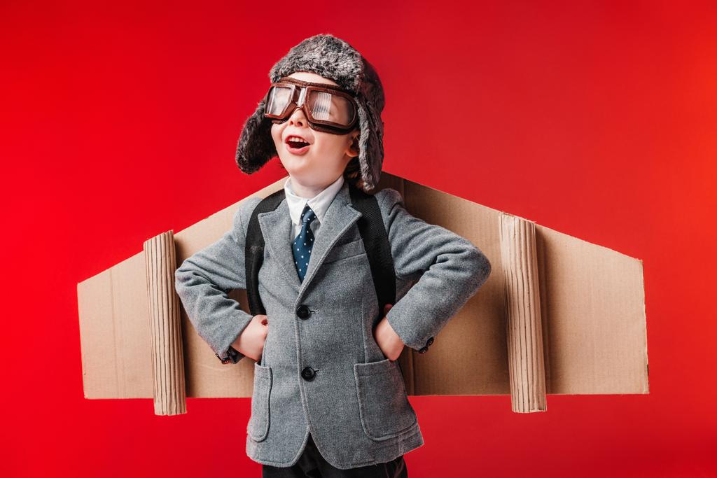 excited boy in suit with paper plane wings and goggles isolated on red - Photo, Image