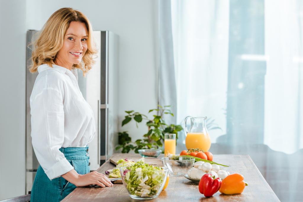 Seitenansicht einer glücklichen erwachsenen Frau, die in die Kamera schaut, während sie in der Küche Salat zubereitet - Foto, Bild