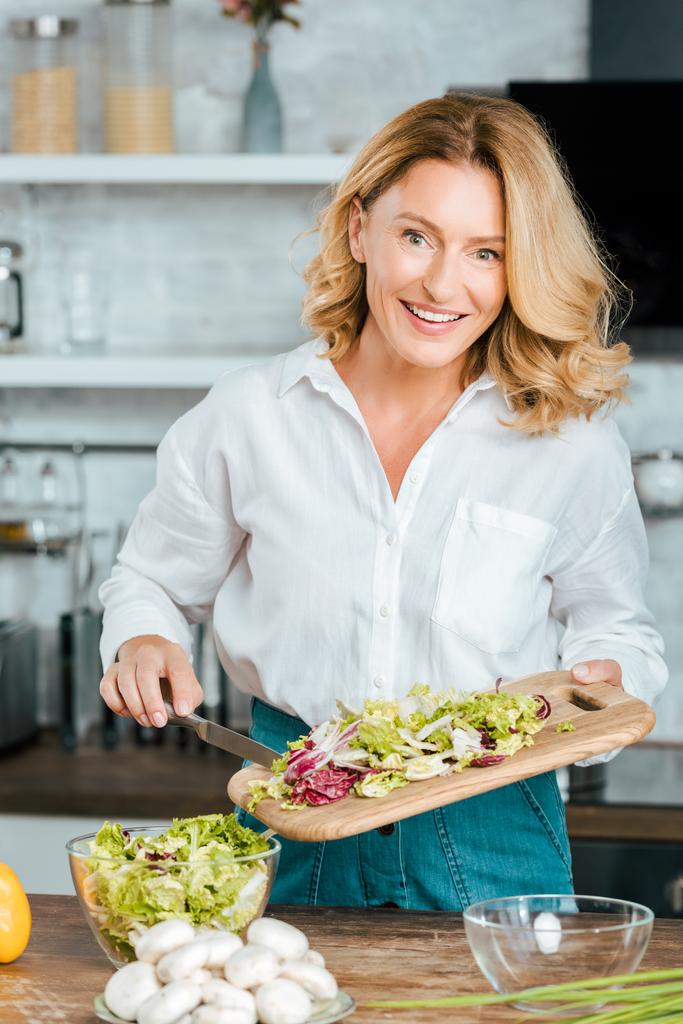 feliz mujer adulta haciendo ensalada saludable en la cocina y mirando a la cámara
 - Foto, Imagen