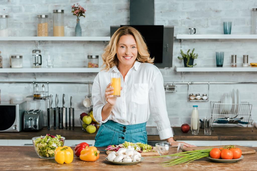 beautiful adult woman drinking orange juice while cooking at kitchen and looking at camera - Photo, Image