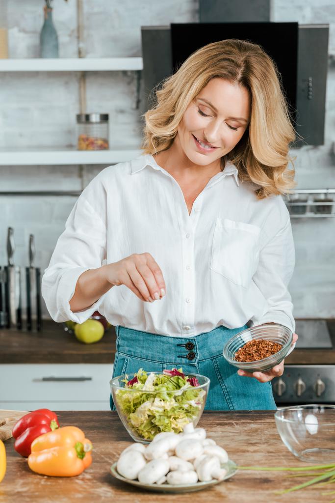 schöne erwachsene Frau verschüttet Gewürz auf Salat in Schüssel in der Küche - Foto, Bild