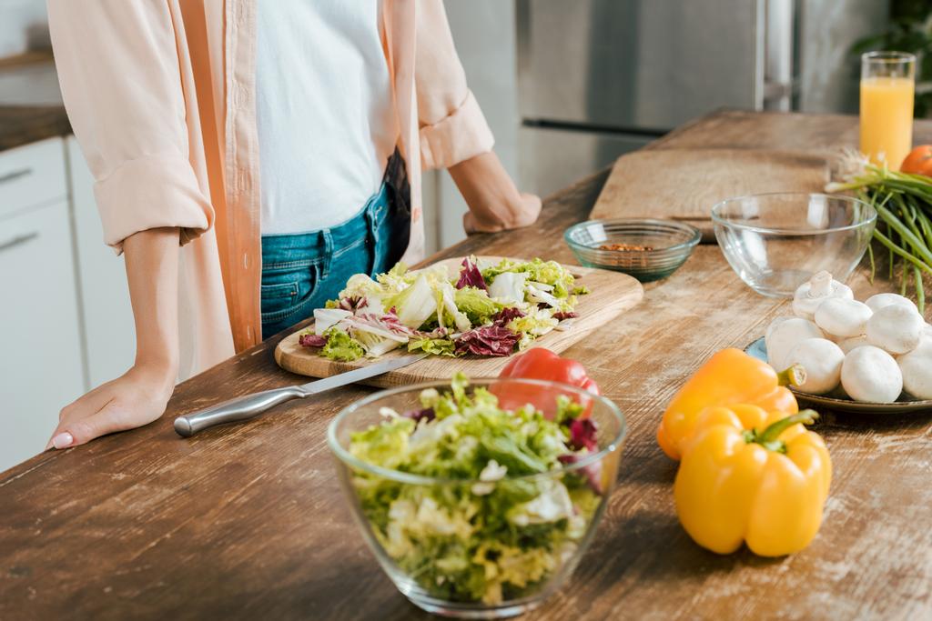 обрізаний знімок жінки, що стоїть біля столу з овочами для салату на кухні
 - Фото, зображення