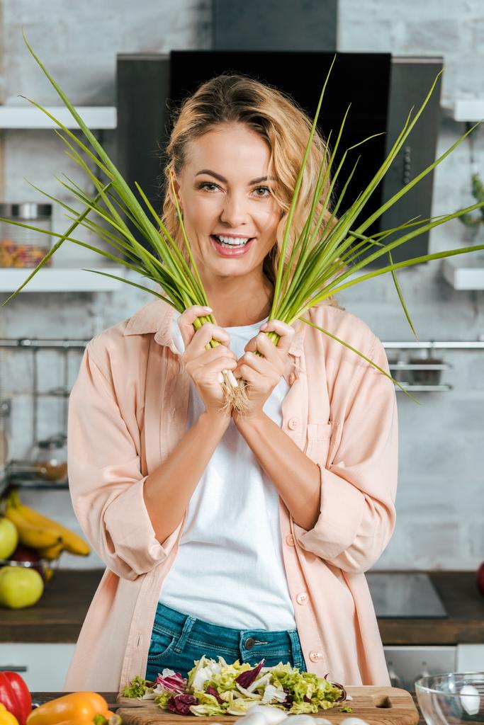 šťastná mladá žena s pórkem, při vaření v kuchyni při pohledu na fotoaparát - Fotografie, Obrázek