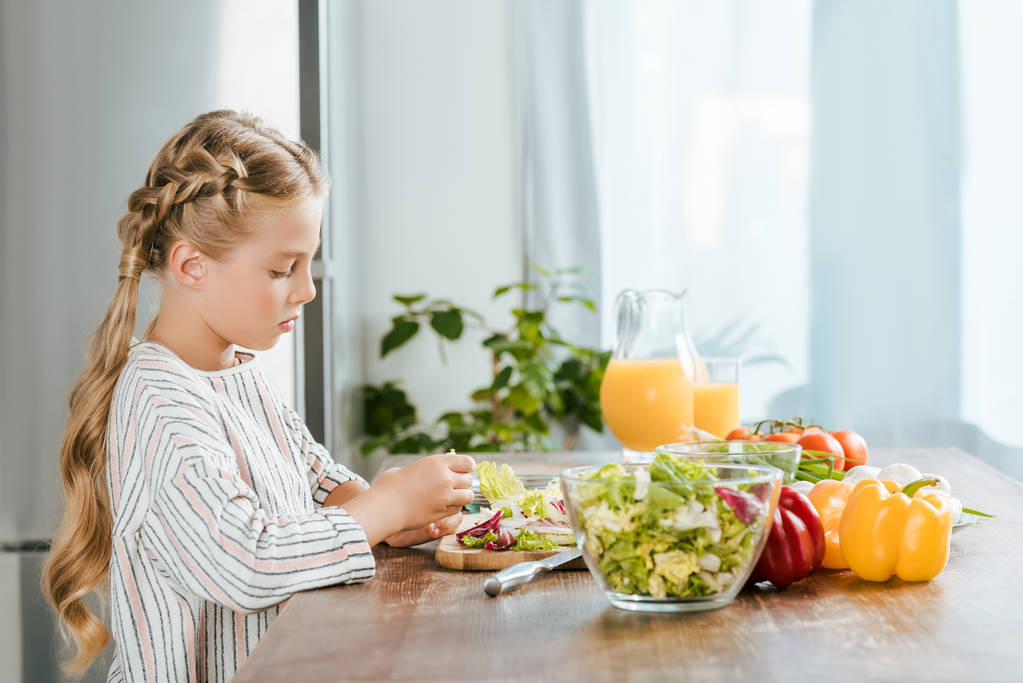 vista lateral da criança pequena concentrada que faz a salada na cozinha
 - Foto, Imagem