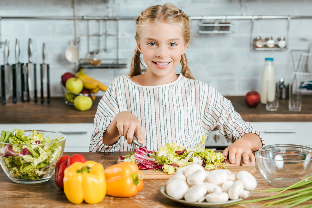 pequena criança feliz cortando alface para salada na cozinha e olhando para a câmera
 - Foto, Imagem