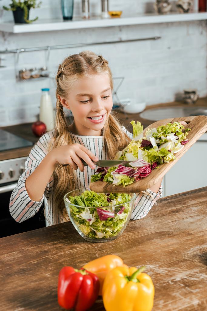 glimlachend kleine kind gieten gesneden sla in kom voor salade op keuken - Foto, afbeelding