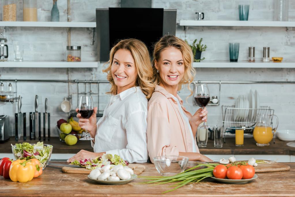 ώριμη μητέρα και κόρη ενηλίκων με ποτήρια κόκκινο κρασί στέκεται πλάτη με πλάτη στην κουζίνα και βλέπουν τα φωτογραφικών μηχανών - Φωτογραφία, εικόνα