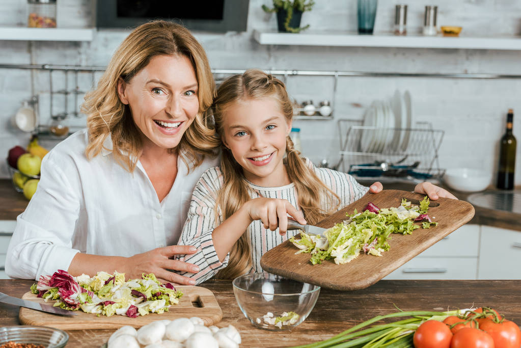 Gelukkig volwassen moeder en dochtertje maken salade samen thuis en kijken naar camera - Foto, afbeelding