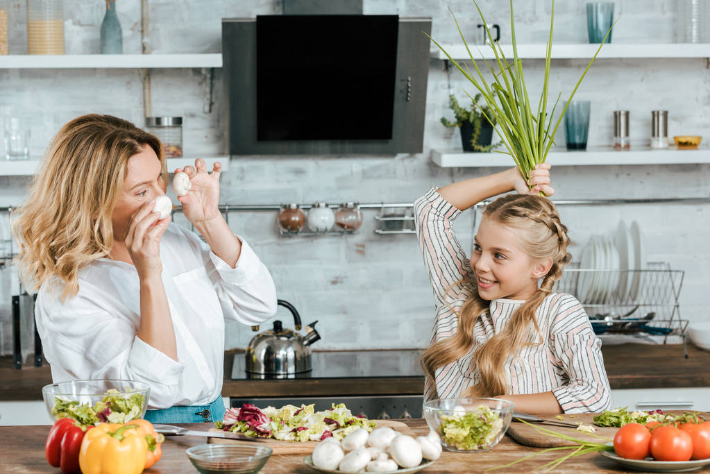sonrientes madre e hija jugando con la comida mientras cocinan mirándose en casa
 - Foto, Imagen