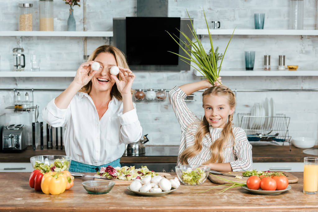 lachende moeder en dochter spelen met voedsel tijdens het koken op de camera thuis kijken - Foto, afbeelding