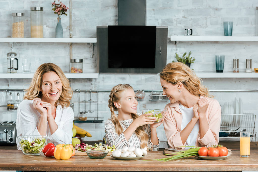 happy little child preparing salad with mother and grandmother at kitchen - Photo, Image