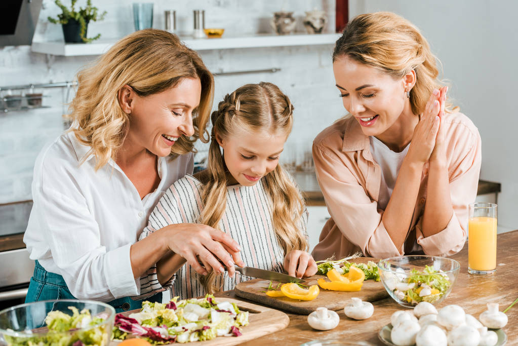 adorable little child cutting vegetables for salad with mother and grandmother at home - Photo, Image