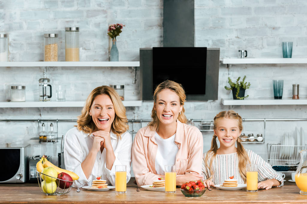 rire trois générations de femmes avec des crêpes et du jus d'orange pour le petit déjeuner à la maison en regardant la caméra
 - Photo, image