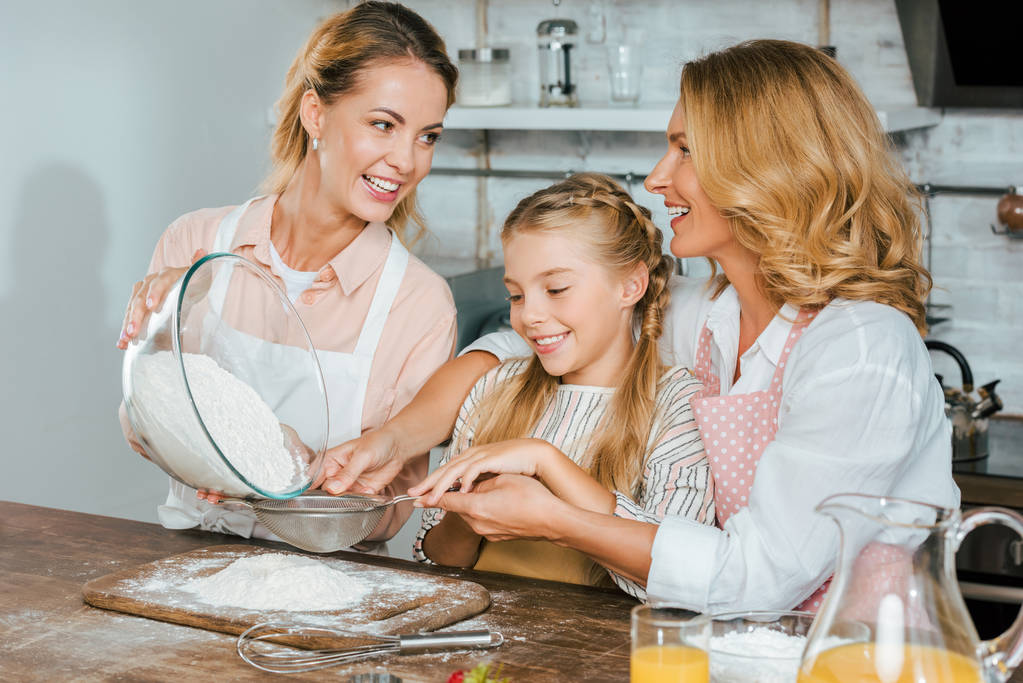 szczęśliwe dziecko z matką i babcią, nalewanie mąkę przez sito podczas wyrabiania ciasta w domu - Zdjęcie, obraz