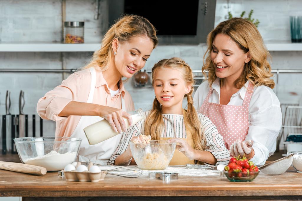 Улыбающийся ребенок с мамой и бабушкой, которые вместе делают тесто дома
 - Фото, изображение