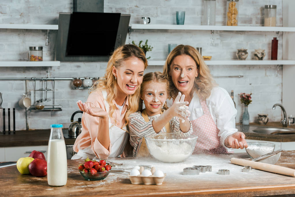 glückliches Kind klatscht mit Mehl in die Hände, während es zu Hause mit Mutter und Großmutter kocht - Foto, Bild