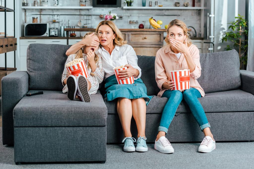 bang kind met moeder en grootmoeder kijken naar film op Bank thuis met emmers van popcorn - Foto, afbeelding