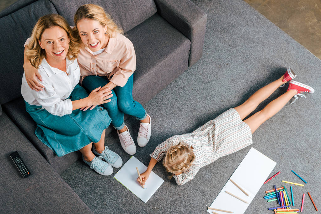 high angle view of happy mother and grandmother sitting on couch while child drawing on floor at home - Photo, Image
