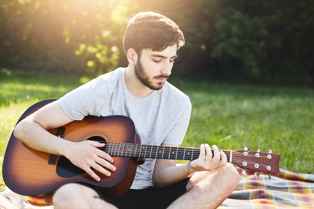 horizontales Porträt eines bärtigen modischen Typen, der mit überkreuzten Beinen sitzt und Gitarre spielt, während er sich im grünen Gras ausruht und die Sonne bewundert. talentierter Musiker singt Lieder und spielt Musikinstrument. - Foto, Bild