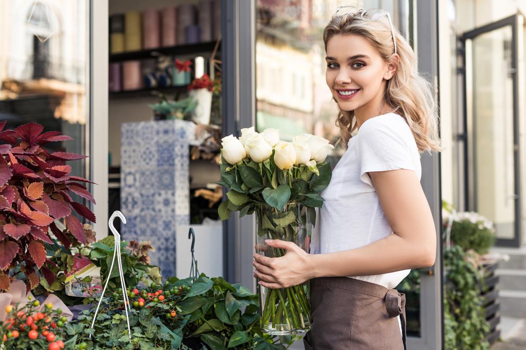 attraente fiorista in possesso di vaso con rose bianche vicino negozio di fiori e guardando la fotocamera
 - Foto, immagini