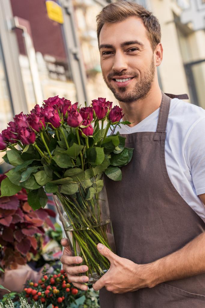 lachende knappe bloemist glazen pot met donkerrood rozen te houden in de buurt van bloemenwinkel - Foto, afbeelding