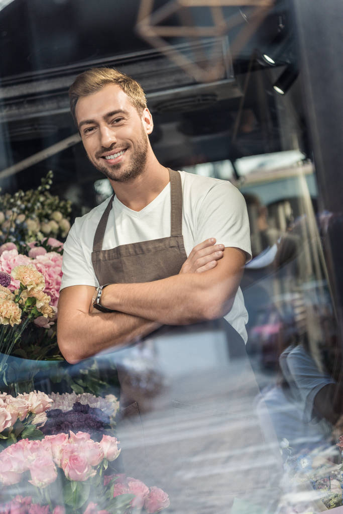 view through window of handsome florist in apron standing with crossed arms in flower shop and looking at camera - Photo, Image