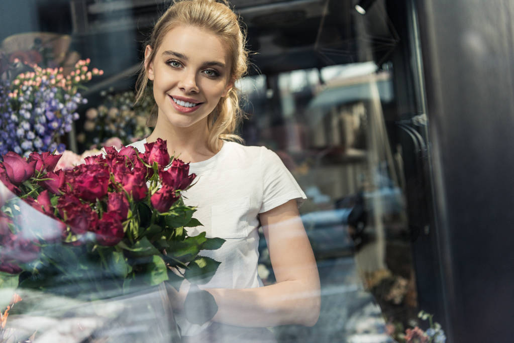 view through window of smiling attractive florist holding bouquet of burgundy roses in flower shop - Photo, Image