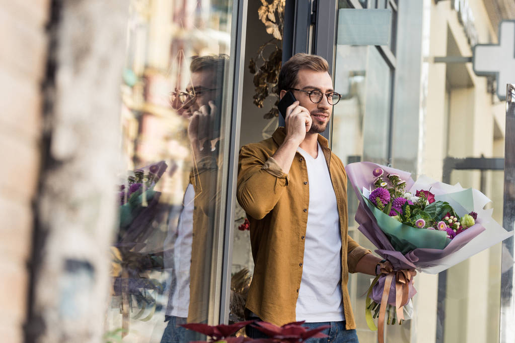 handsome man going out from flower shop with bouquet and talking by smartphone - Photo, Image