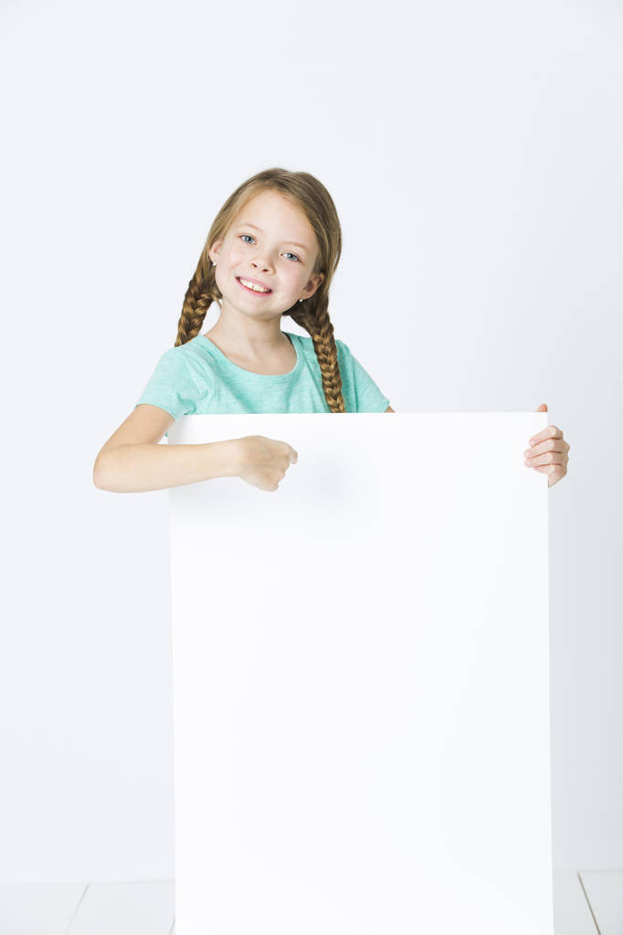 chica feliz con trenzas en camiseta turquesa sonriendo y apuntando a tablero vacío aislado sobre fondo blanco
 - Foto, Imagen