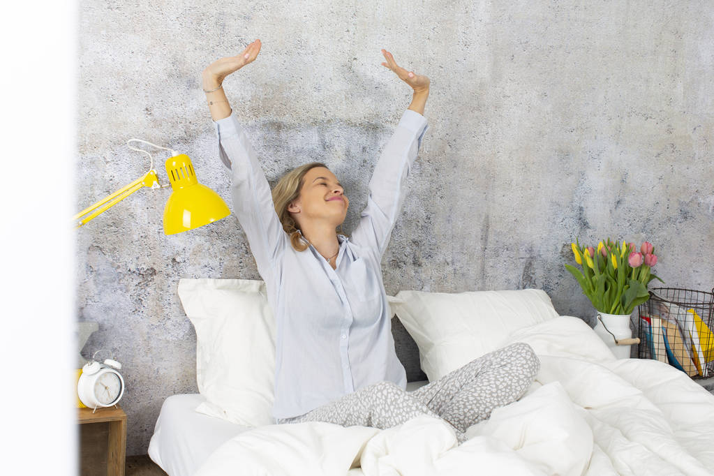 zufriedene junge Frau im Schlafanzug lächelt, während sie morgens auf dem Bett sitzt - Foto, Bild