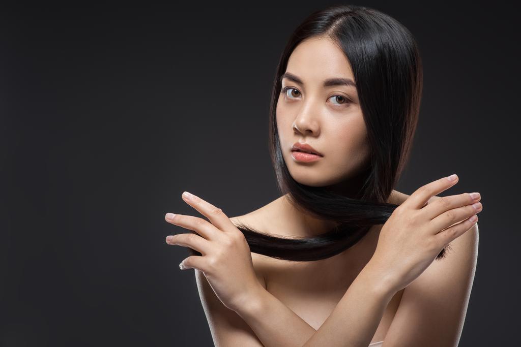 портрет молодой азиатской женщины с красивыми и здоровыми темными волосами, смотрящей на камеру, изолированную на черном
 - Фото, изображение