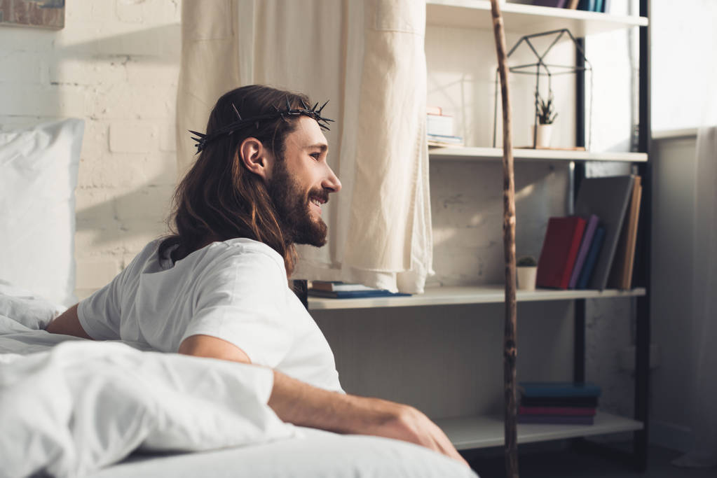 вид сбоку улыбающегося Иисуса, сидящего на полу возле кровати в спальне дома в утреннее время
  - Фото, изображение