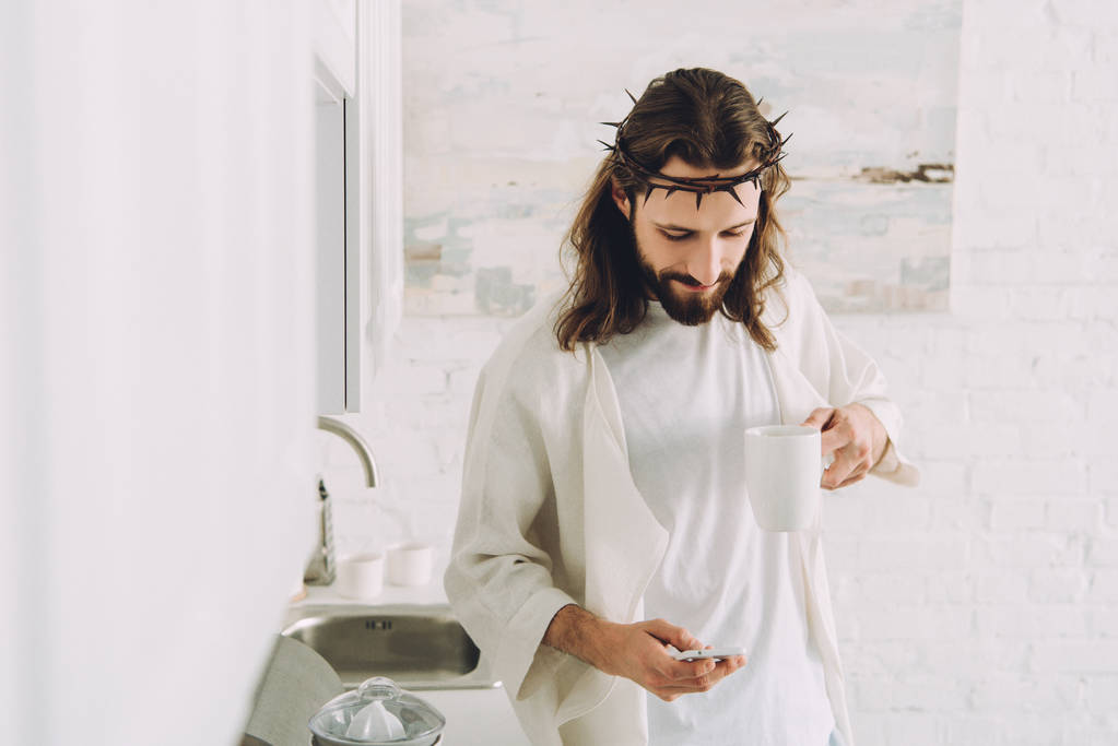 Jesus feliz na coroa de espinhos de pé com xícara de café e usando smartphone na cozinha em casa
  - Foto, Imagem