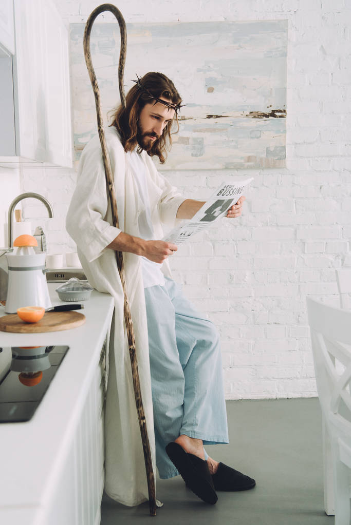 focused Jesus with wooden staff reading business newspaper in kitchen at home - Photo, Image