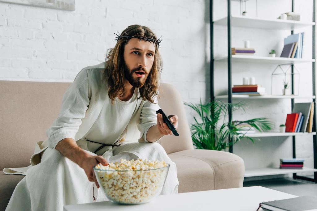 σοκαρισμένος ο Ιησούς με ακάνθινο στεφάνι, βλέποντας τηλεόραση και καθιστικό με μπολ από ποπ-κορν στον καναπέ στο σπίτι - Φωτογραφία, εικόνα