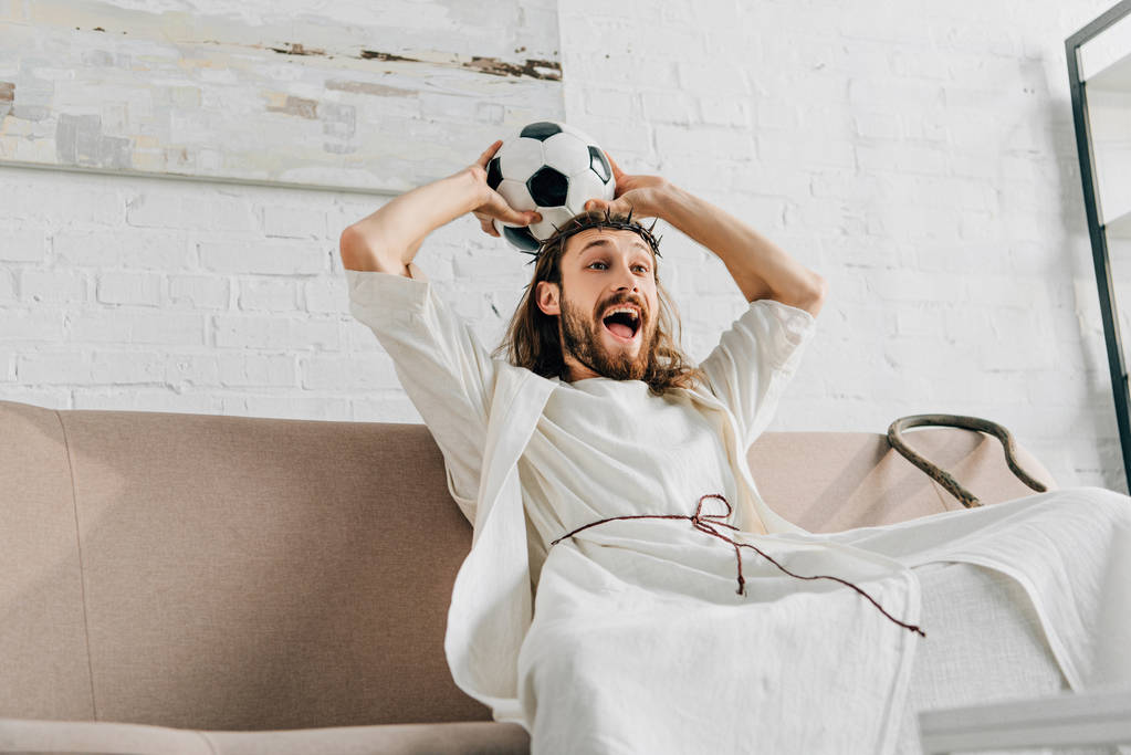 Jesus animado na coroa de espinhos segurando bola de futebol acima da cabeça e assistindo jogo de futebol em casa
 - Foto, Imagem