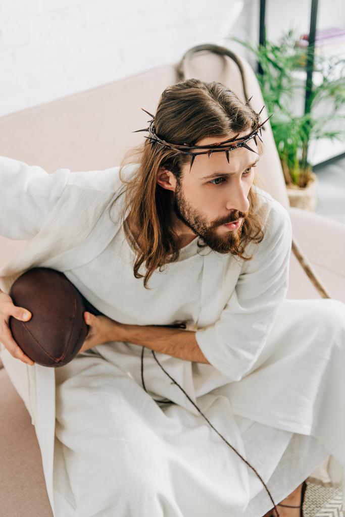 wysoki kąt widzenia koncentruje się Jezusa w koronie cierniowej, trzymając piłkę i oglądania futbolu amerykańskiego na kanapie w domu - Zdjęcie, obraz