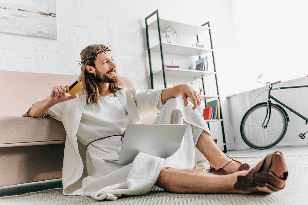 Jesús sonriente sentado en el suelo con tarjeta de crédito y haciendo compras en línea en el ordenador portátil cerca del sofá en casa
 - Foto, imagen