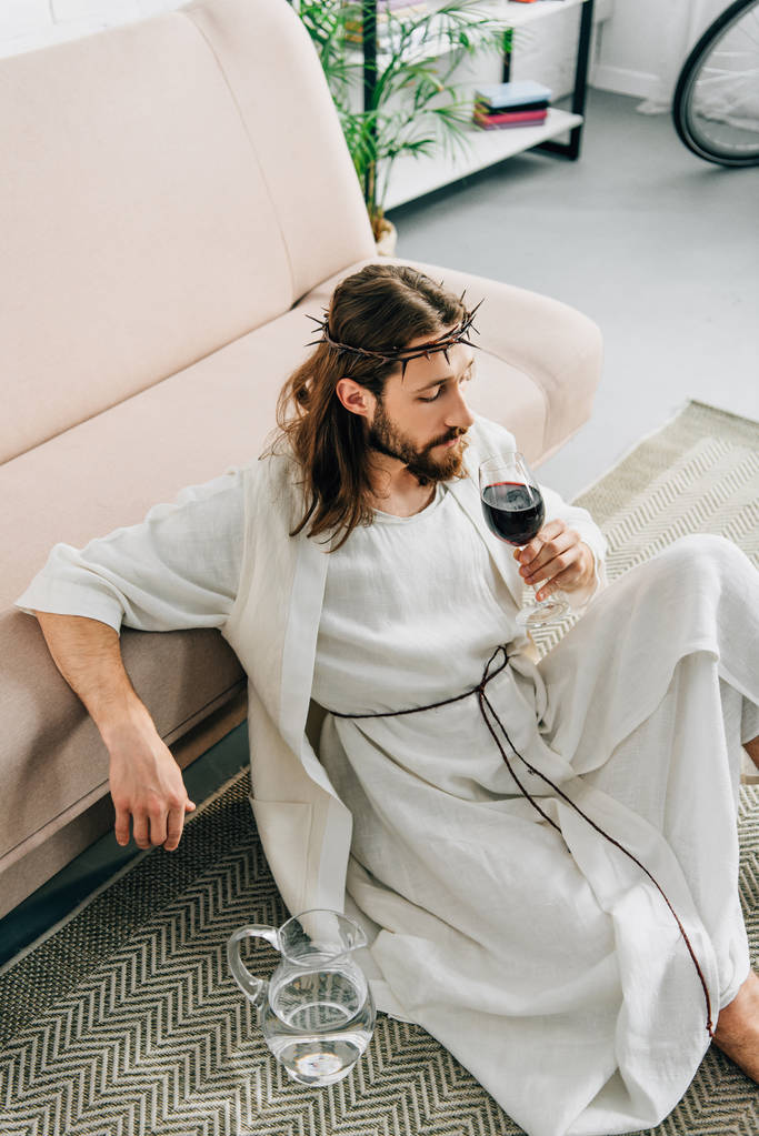 vista de alto ângulo de Jesus em coroas de espinhos e roupão sentado no chão perto jarro de água e beber vinho de vidro em casa
 - Foto, Imagem