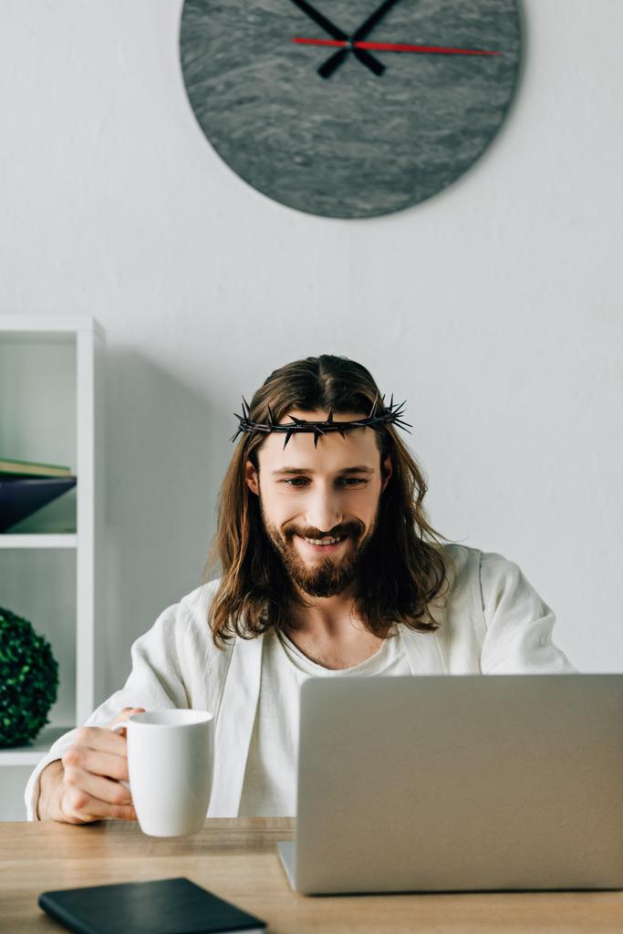 cheerful Jesus in crown of thorns with coffee cup using laptop at table in modern office - Photo, Image