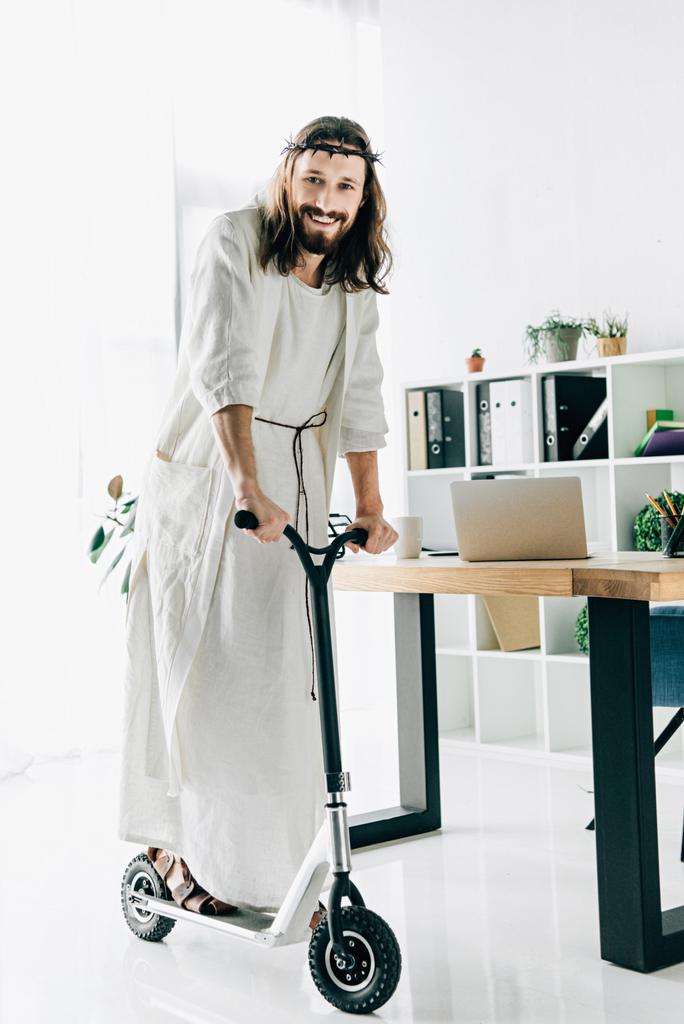 happy Jesus in crown of thorns and robe riding on kick scooter in modern office  - Photo, Image