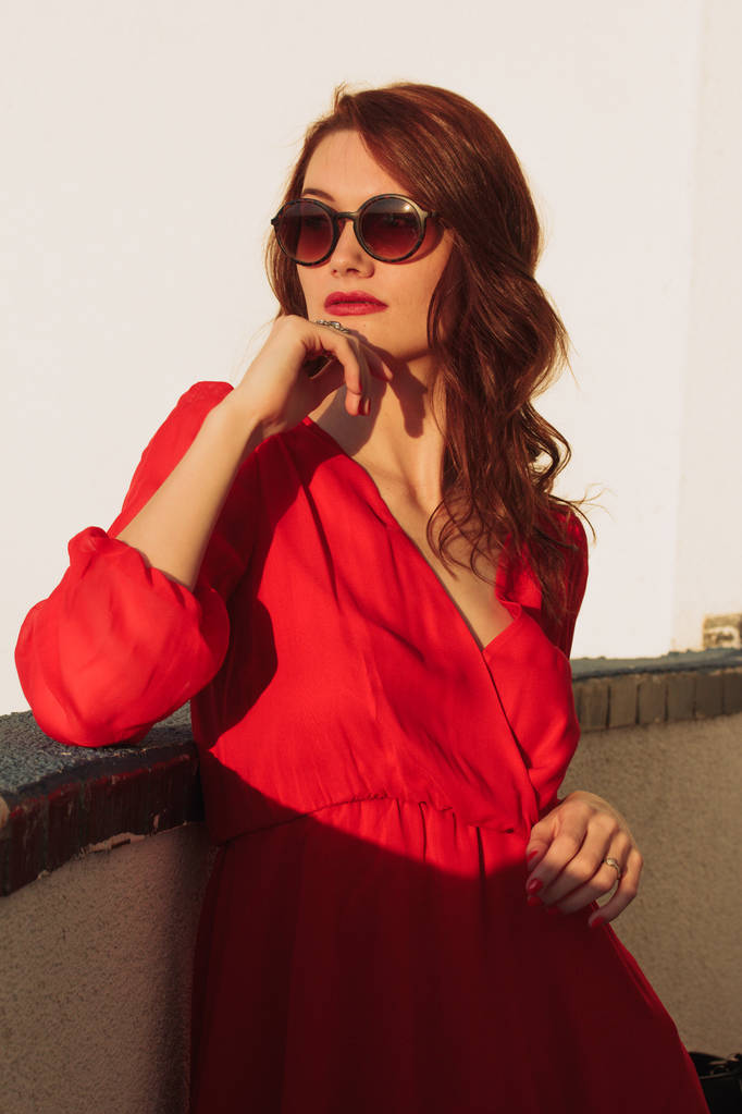 Giovane ragazza bruna in piedi sul balcone al tramonto in un vestito rosso e occhiali da sole
 - Foto, immagini