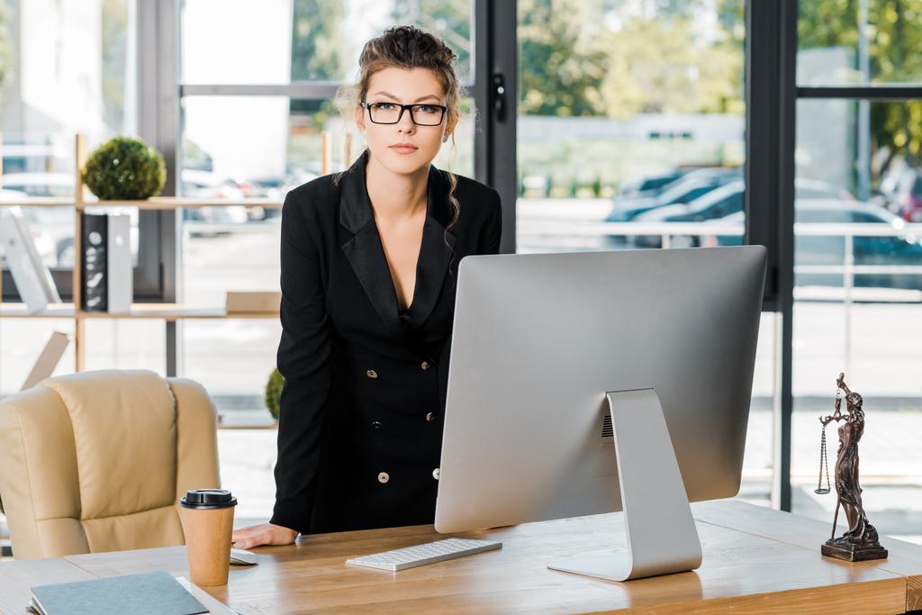 attractive businesswoman in glasses leaning on table with computer and looking at camera in office - Photo, Image