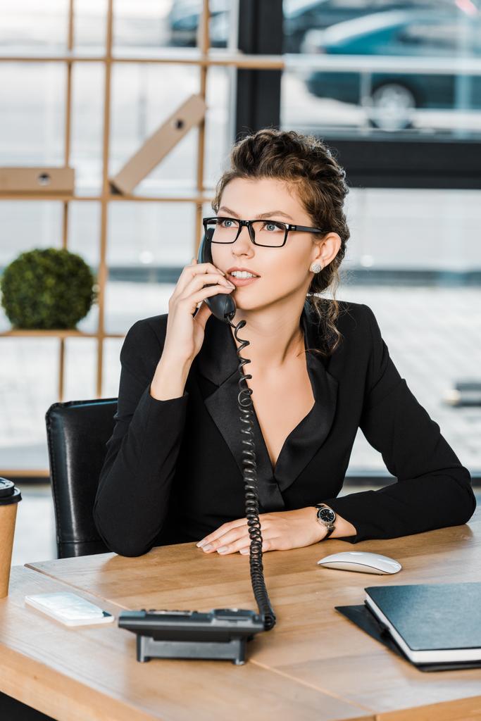 привлекательная деловая женщина разговаривает по стационарному телефону в офисе и смотрит в сторону
 - Фото, изображение