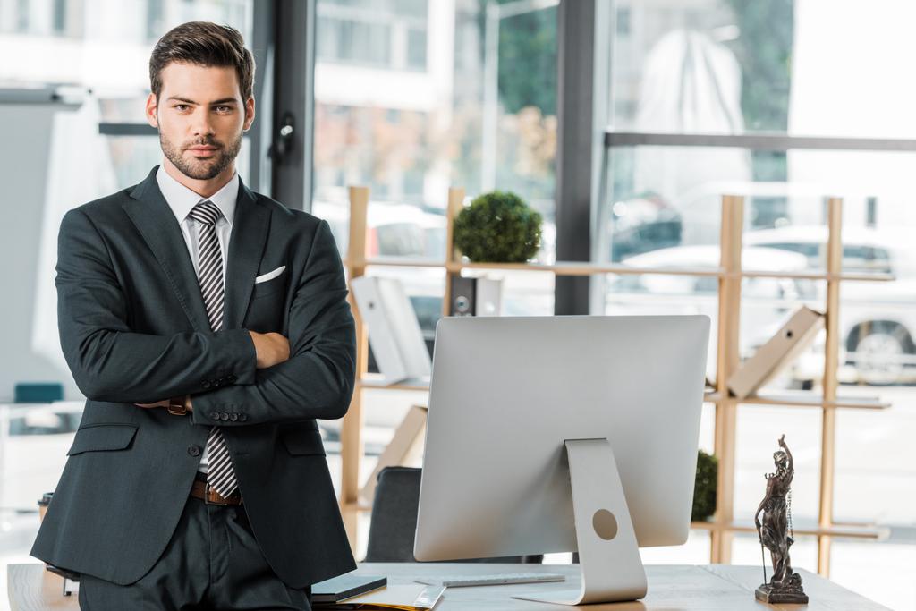 handsome businessman standing with crossed arms near table in office - Photo, Image