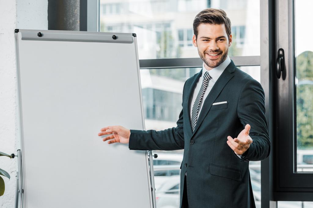 Porträt eines gut gelaunten Geschäftsmannes im Anzug, der an einer leeren weißen Tafel im Büro steht - Foto, Bild